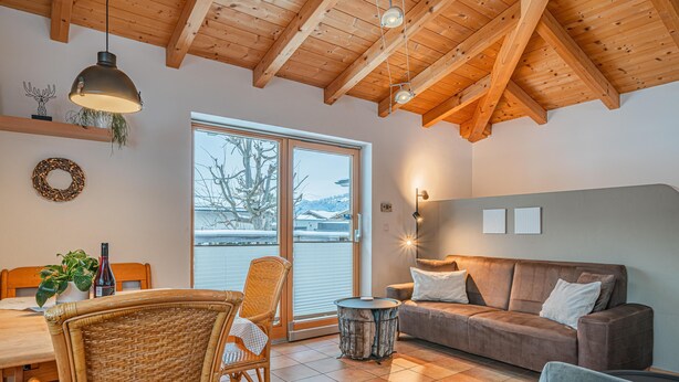 A cozy living room with a wooden ceiling, a brown sofa, a grey armchair, and a dining area with wicker chairs and a table. - Detached holiday home in centre of Mittersill (Mittersill)