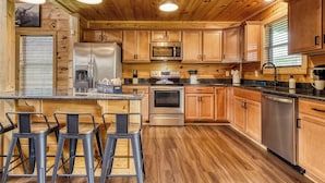 A modern kitchen with wooden cabinets, stainless steel appliances, and a bar area with stools. - TWO Cabins!  Sleeps 24, Themed Bunk Rooms for The Kids, Hot Tubs, Mountain Views (Sevierville)