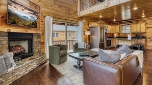 A cozy cabin interior with a stone fireplace, a dining area, a kitchen, and a balcony with a view. - TWO Cabins!  Sleeps 24, Themed Bunk Rooms for The Kids, Hot Tubs, Mountain Views (Sevierville)