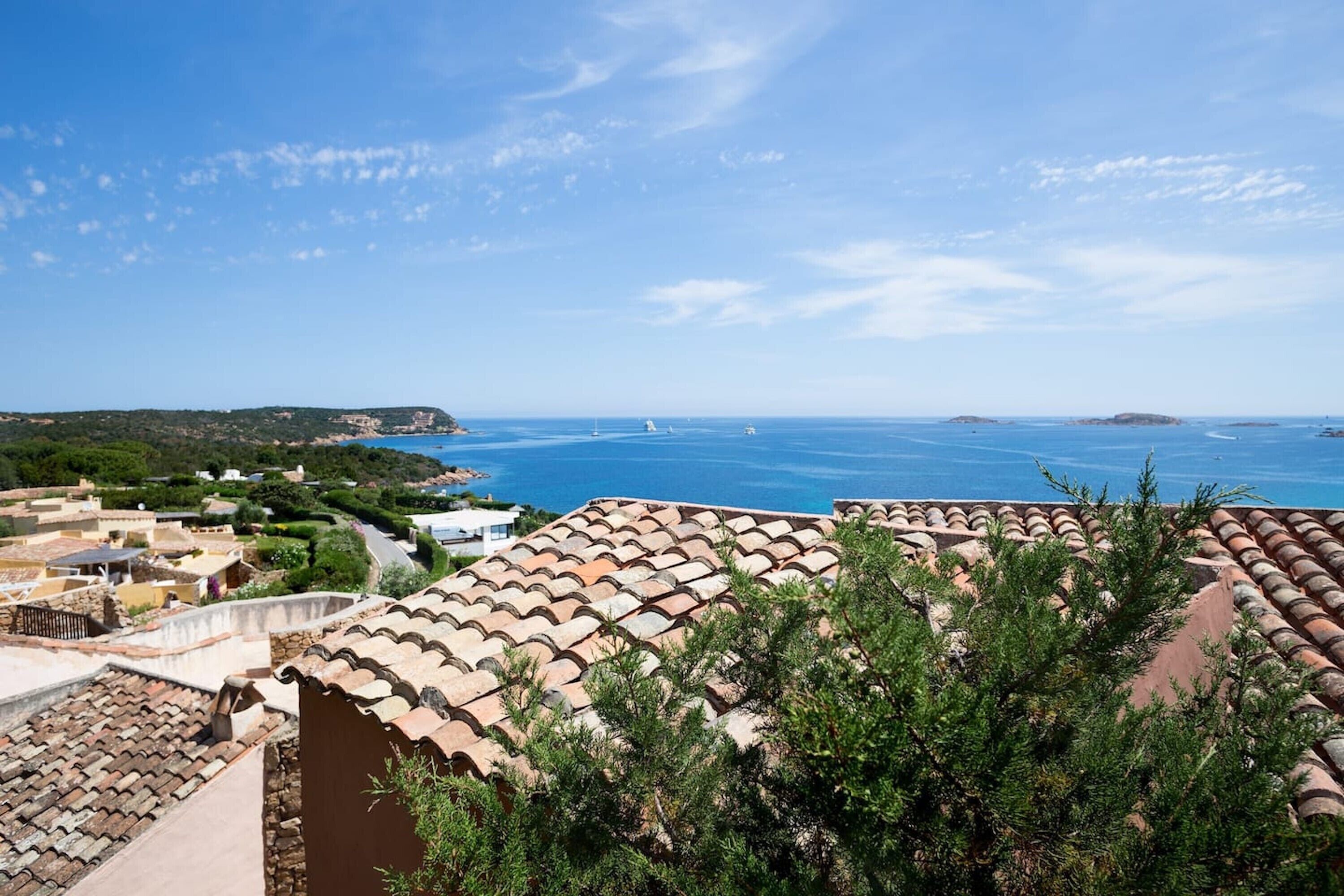 A coastal view with terracotta rooftops, a clear sky, and distant boats.