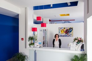 Un mostrador de recepción con una persona detrás, un cuadro en la pared y una planta sobre el mostrador.
