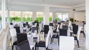 Un comedor espacioso con varias mesas y sillas, amplias ventanas que dan a un jardín, y una cocina visible al fondo.