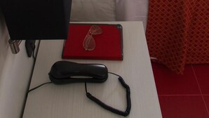 Habitación de hotel con una cama individual, una mesita de noche con lámpara, un teléfono y una carpeta roja.