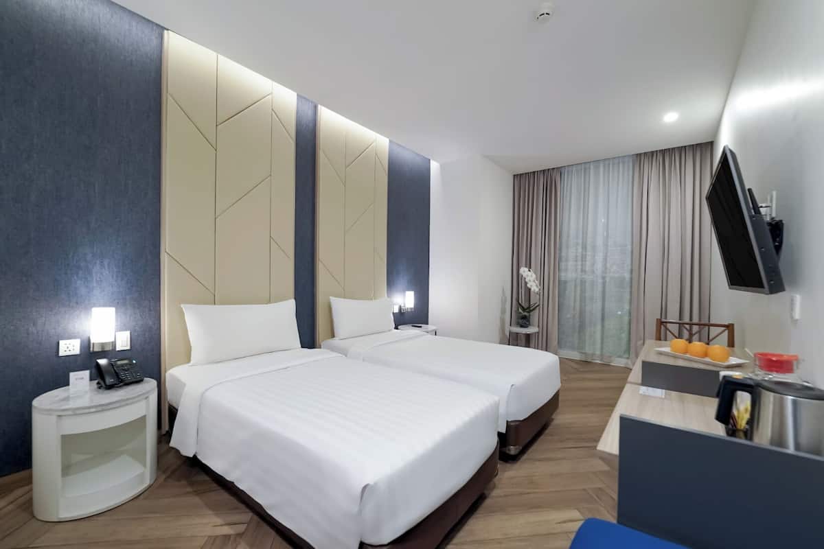 A hotel room with two beds, a desk with a chair, a TV mounted on the wall, and a window with curtains.