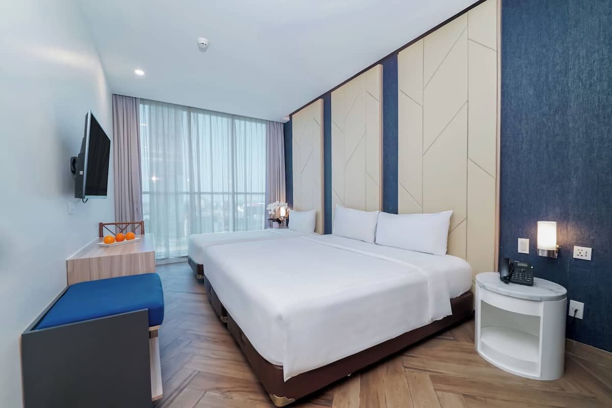 A hotel room with a large bed, a desk with a chair, a flat-screen TV, a bedside table with a phone, and a window with curtains.
