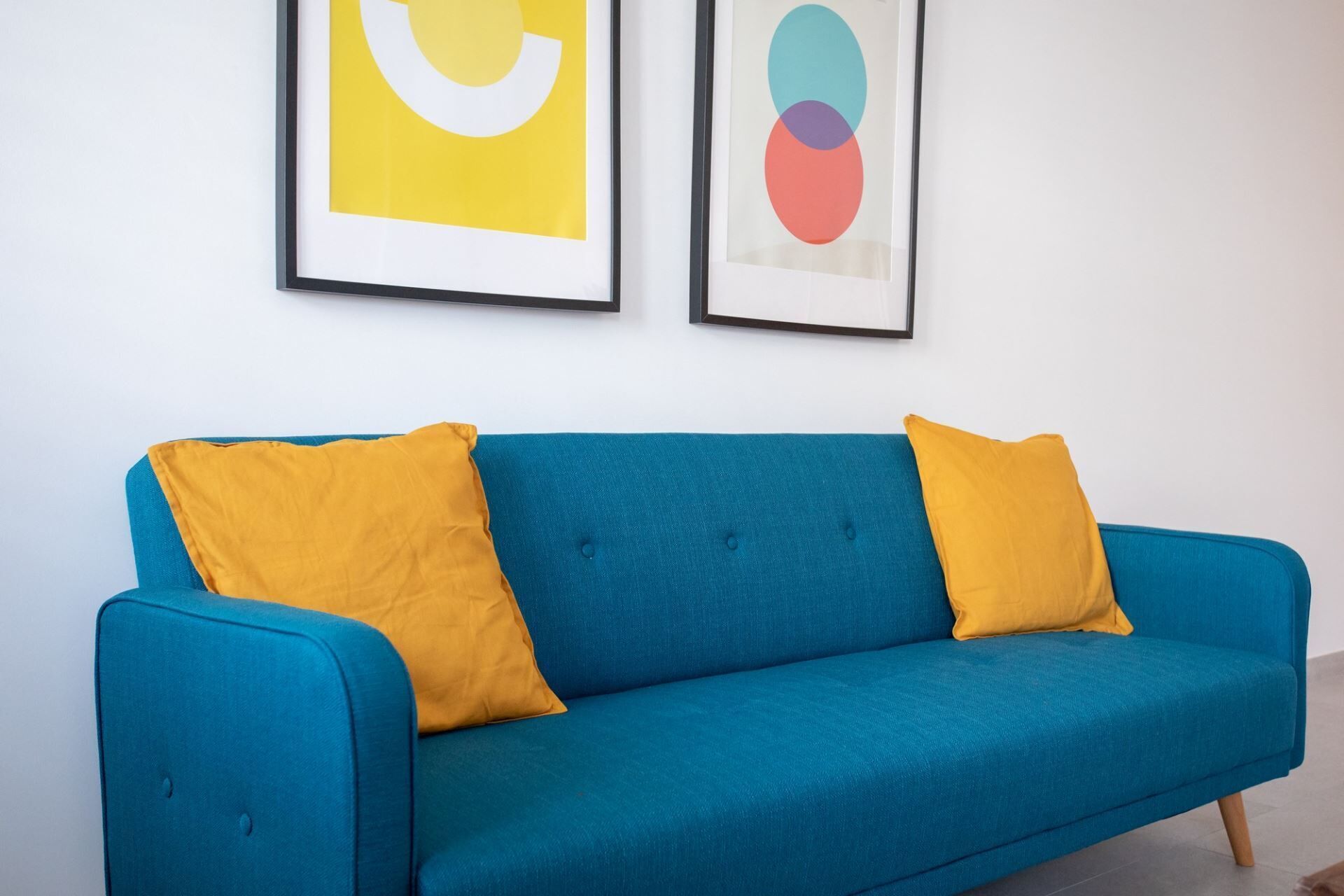 A teal sofa with two mustard yellow cushions against a white wall, flanked by two framed abstract artworks.