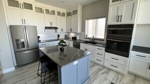 A modern kitchen with white cabinetry, a central island, a double oven, and a stainless steel refrigerator. - Lake View Modern House with Beautiful Sunsets! (Page)