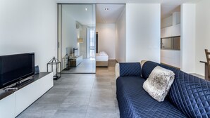 A modern living room with a flat-screen TV, a sofa with a quilted cover, and a glass partition leading to a bedroom. - San Maurilio  Apartment (Milano)