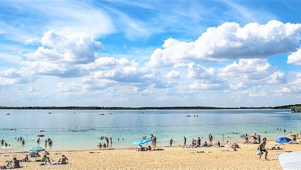 Ein Strand mit badenden und sonnenbächelnden Menschen, einem klaren Himmel und Booten auf dem Wasser.