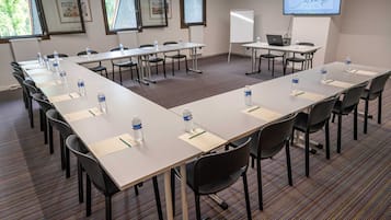 Un salón de conferencias con mesas largas, sillas, botellas de agua y una pantalla de proyector mostrando un logotipo.