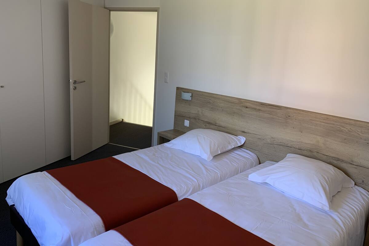 A hotel room with two beds, a wooden headboard, a door, and a view of a corridor.