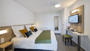 Ein modernes Hotelzimmer mit einem ordentlich bezogenen Bett, einem Nachttisch, einem Sessel, einem an der Wand montierten Fernseher und einer kleinen Küchenzeile.