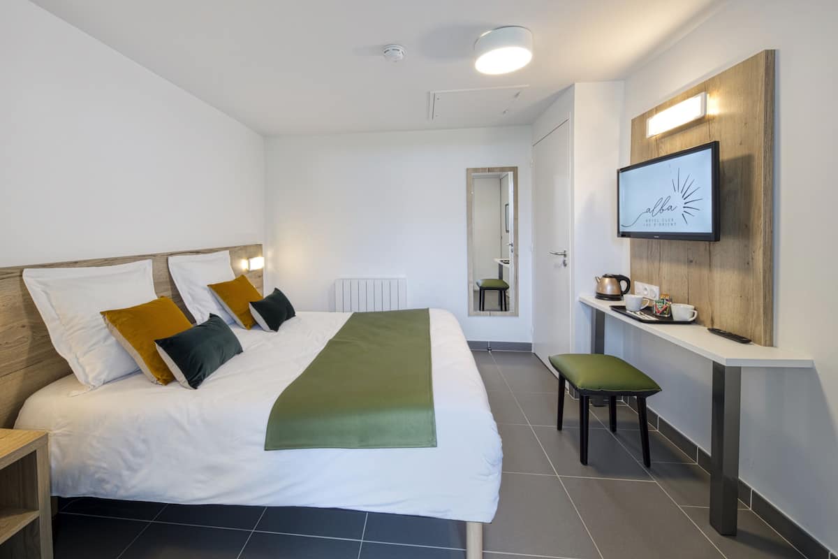A modern hotel room with a neatly made bed, a bedside table, a chair, a TV mounted on the wall, and a small kitchenette area.