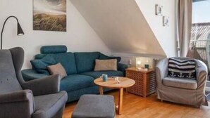 A cozy loft-style living room with a sloped ceiling, a teal sofa, a grey armchair, a small wooden coffee table, and a grey ottoman. - Waldstr. 34 Wo.05 - Wo.05 (Kühlungsborn)