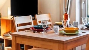 A dining area with a wooden table set for two, a television in the background, and a window with curtains. - Waldstr. 34 Wo.05 - Wo.05 (Kühlungsborn)