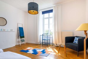Ein Zimmer mit einem Bett, einem Schreibtisch mit Stuhl, einem Sofa, einem Fenster mit Vorhängen, einer Pendelleuchte und einer Heizung.