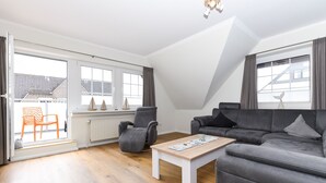A modern living room with a grey sofa, a white coffee table, and a grey armchair. There is a window with curtains and a view of buildings outside. - Ferienwohnung Juist - Ferienwohnungen Min Huus (Neuharlingersiel)