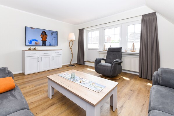 A modern living room with a flat-screen TV, a wooden coffee table, grey sofas, a grey armchair, and a window with curtains.
