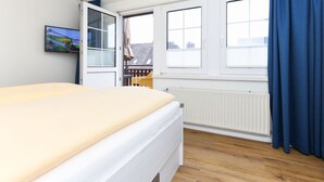 A bedroom with a bed, a window with blinds, a radiator, and a TV on the wall. - Ferienwohnung Norderney - Ferienwohnungen Min Huus (Neuharlingersiel)