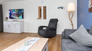 A modern living room with a wooden coffee table, a grey sofa, a black leather chair, a television, and a lamp. - Ferienwohnung Norderney - Ferienwohnungen Min Huus (Neuharlingersiel)