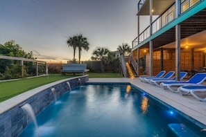 A swimming pool with a waterfall feature, surrounded by a wooden deck with lounge chairs and a covered area with stairs leading to an upper level. - Exquisite Oceanfront Oasis: Immerse in Luxury with Pool & Putting Green at this (Folly Beach)