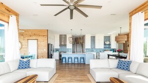 Ein modernes Wohnzimmer mit einem großen Deckenventilator, eine Küche mit Kochinsel und ein Essbereich mit blauen Stühlen.