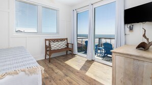 A room with a bed, a wooden chair, a view of the beach through a sliding glass door, and a small table with a decorative item. - Exquisite Oceanfront Oasis: Immerse in Luxury with Pool & Putting Green at this (Folly Beach)