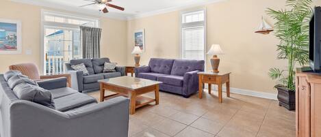 A spacious living room with two sofas, a coffee table, and a TV stand. There are ceiling fans, a potted plant, and artwork on the walls.