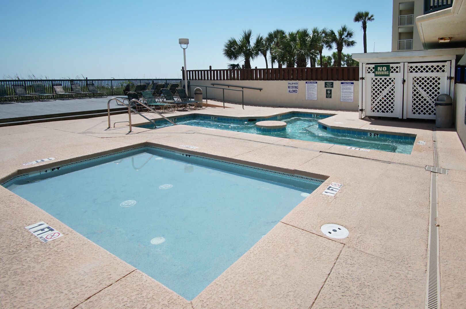 Un espace piscine comprenant plusieurs bassins, un poste de surveillance pour les sauveteurs et une terrasse clôturée avec des chaises longues.