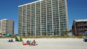 Un hotel moderno de gran altura con varios balcones, una playa arenosa al frente y palmeras.