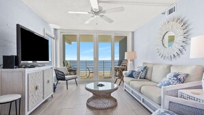 A spacious living room with a large TV, comfortable seating, a ceiling fan, and a view of the ocean through large sliding doors. - Direct ocean front condo with stunning renovations & updated "coastal chic" decor! (North Myrtle Beach)