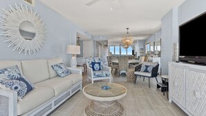 A spacious living room with a large mirror, a ceiling fan, and a dining area with wicker furniture. - Direct ocean front condo with stunning renovations & updated "coastal chic" decor! (North Myrtle Beach)