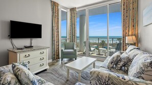 A spacious living room with a large flat-screen TV, a white cabinet with drawers, a sofa set with floral upholstery, a coffee table, and a balcony with a view of the ocean. - Oceanfront luxury condo w/pools, lazy river, fitness center, sky deck and more (North Myrtle Beach)