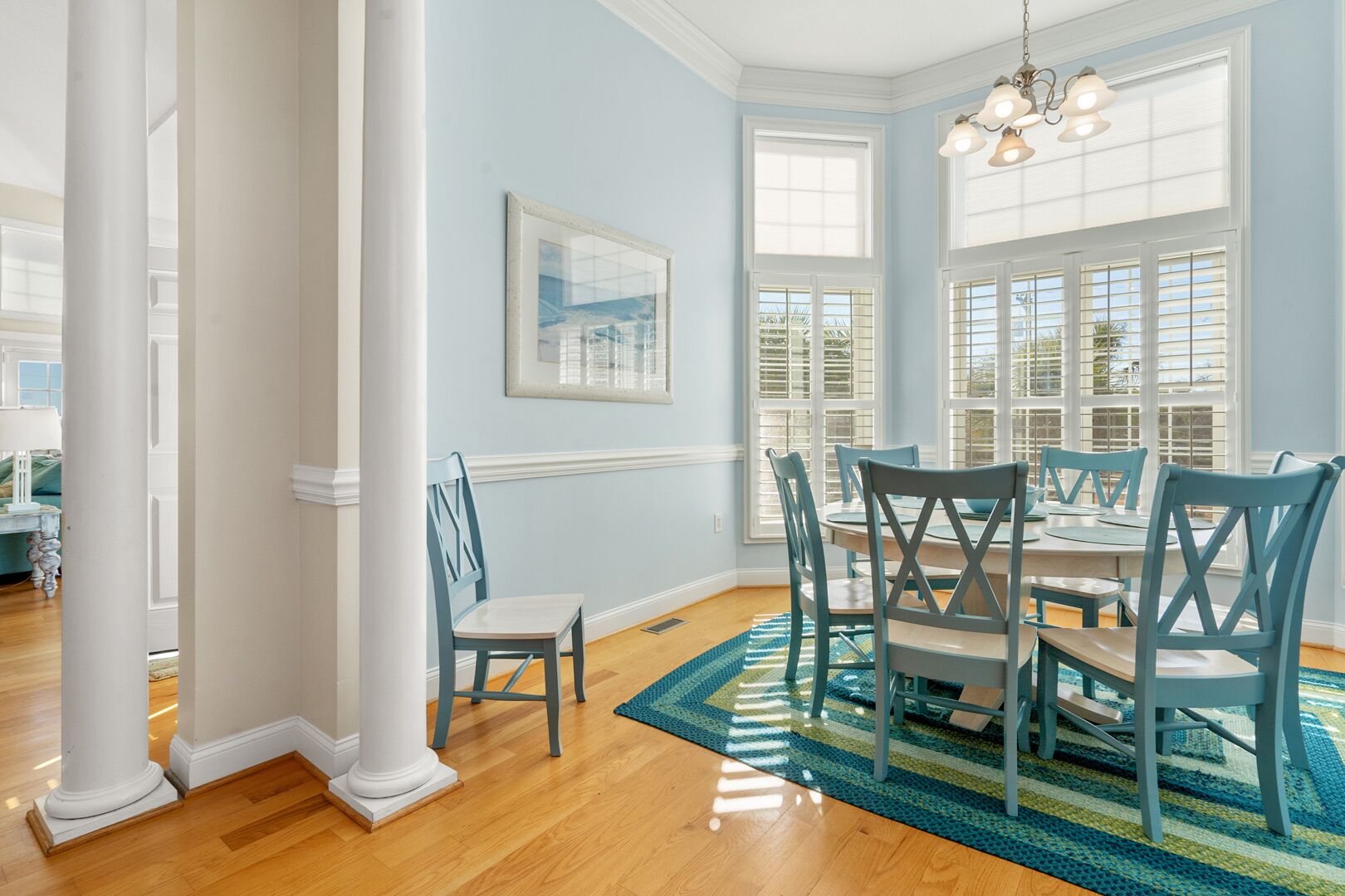 A dining room with a round table, six chairs, a blue and green striped rug, and a framed picture on the wall.