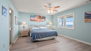 A bedroom with a bed, two nightstands, a ceiling fan, a window with a view, and artwork on the walls. - Luxurious oceanfront home with private pool, putting green, private beach access and more (North Myrtle Beach)