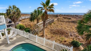 A pool area with a white fence, palm trees, and a view of the beach. - Luxurious oceanfront home with private pool, putting green, private beach access and more (North Myrtle Beach)