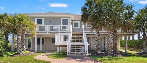 A two-story beachfront property with a balcony, palm trees, and a clear sky.