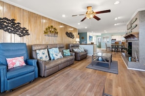 A living room with a blue armchair, two brown sofas, a glass coffee table, wooden flooring, a ceiling fan, and a kitchen in the background. - Immaculate Oceanfront Home - Completely Renovated, Private Pool, Game Room - in Ocean Drive (North Myrtle Beach)