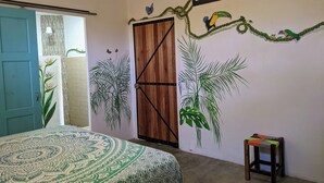 Un dormitorio con cama, una puerta, un taburete pequeño y decoración en la pared, incluyendo un tucán y plantas.