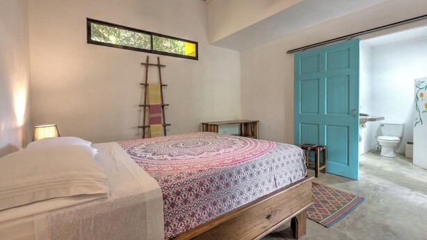 Un dormitorio con cama, una escalera, una mesita pequeña y un baño con inodoro y puerta azul.