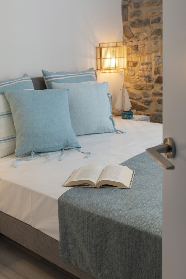 Una cama bien hecha con almohadas azules, un libro sobre la cama y una pared de piedra al fondo.