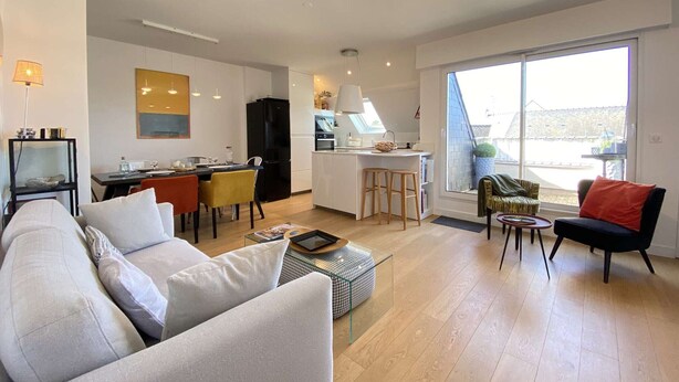 A modern living room with a sofa, a dining area with chairs, a kitchen with a fridge, and a large window offering a view.