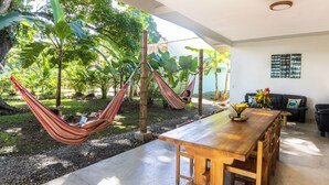 Une table à manger en bois avec des chaises, un hamac et une plante en pot, dans une pièce baignée de lumière et offrant une vue sur un jardin.