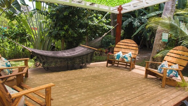 Une terrasse en bois avec un hamac, deux chaises en bois avec des coussins et un banc en bois.