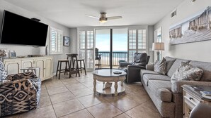 A spacious living room with a large TV, comfortable seating, a dining area, and a view of the ocean through large windows. - Oceanfront 4BR corner condo with balcony views, pools, lazy river, steps to Main Street in NMB. (North Myrtle Beach)