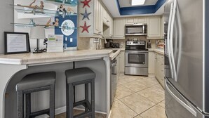 A compact kitchen with a microwave, oven, and stainless steel refrigerator. There are two bar stools and a glass display shelf with decorative items. - Oceanfront 4BR corner condo with balcony views, pools, lazy river, steps to Main Street in NMB. (North Myrtle Beach)