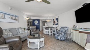A modern living room with a leather sofa, a round coffee table, and a dining area with white chairs and a blue patterned chair. - Oceanfront 4BR corner condo with balcony views, pools, lazy river, steps to Main Street in NMB. (North Myrtle Beach)