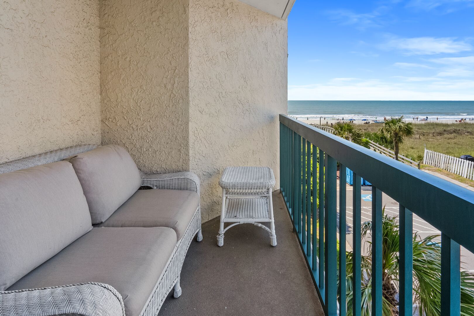 A balcony with a beige sofa and a wicker chair, overlooking a beach with a clear sky.