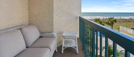 A balcony with a beige sofa and a wicker chair, overlooking a beach with a clear sky.
