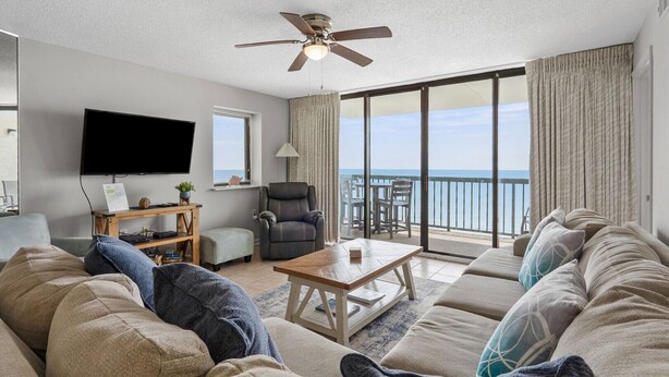 En stue med sofa, sofabord, fjernsyn og en lænestol. Der er en balkon med udsigt over havet.